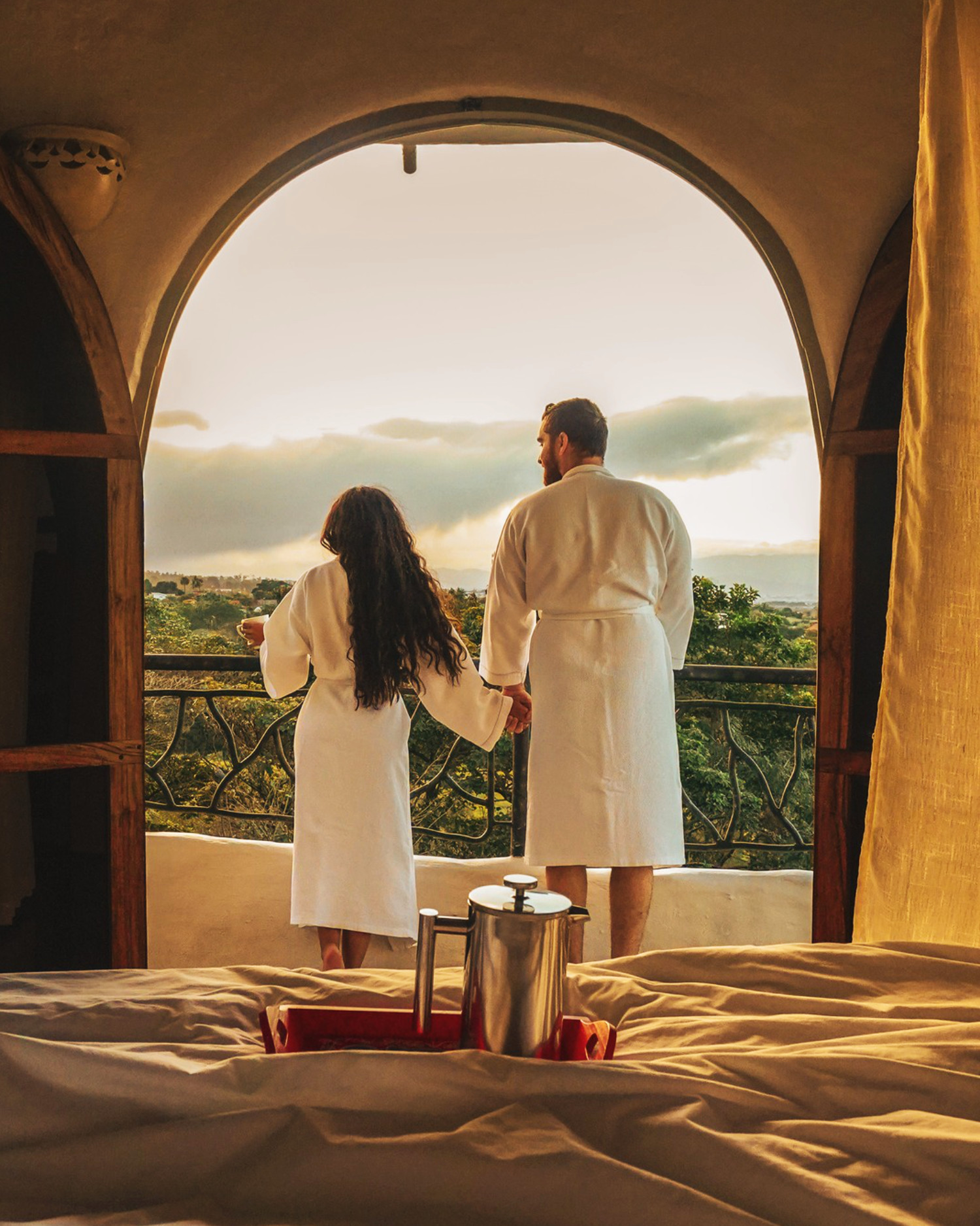 Two guests in white robes standing at an arched window overlooking lush greenery at Finca Rosa Blanca